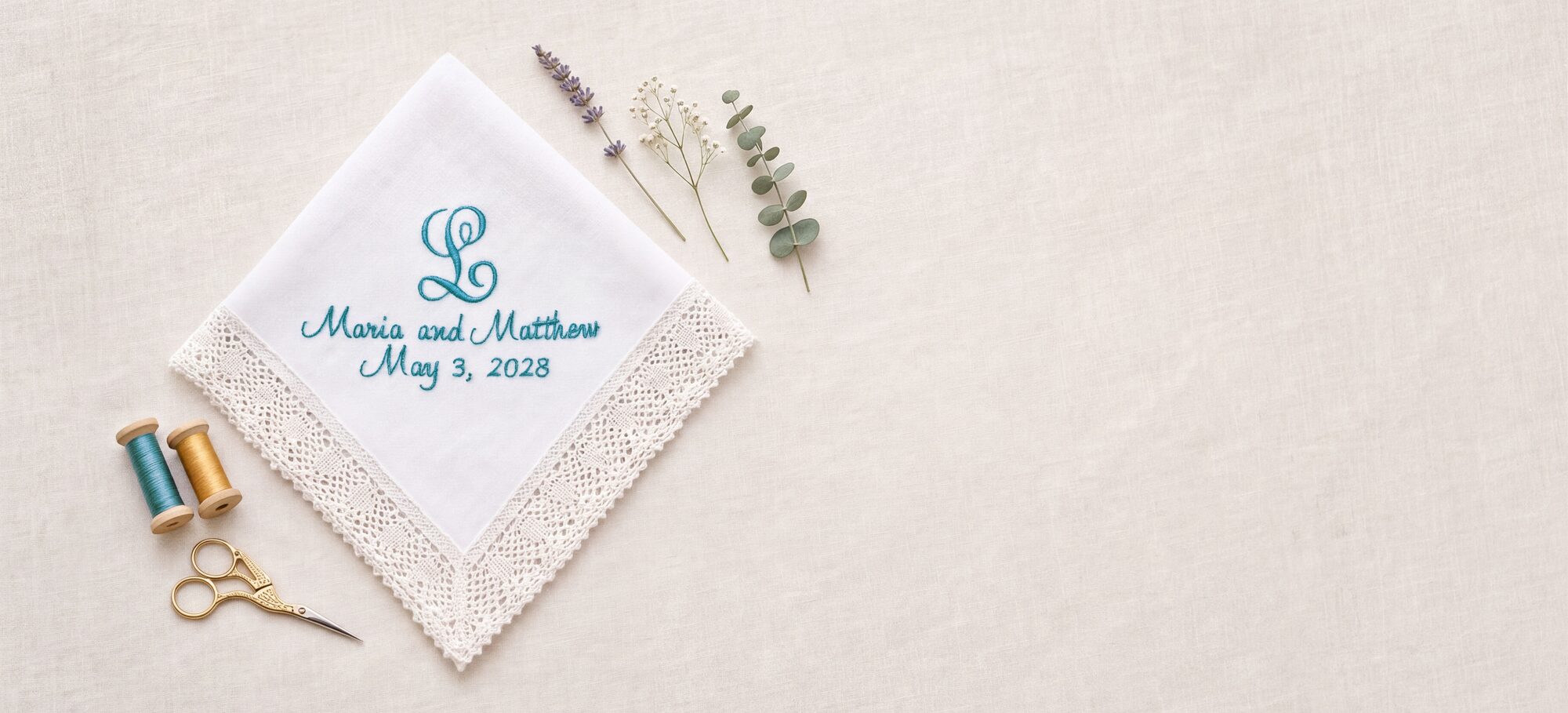 A white cotton handkerchief with a crochet lace border, personalized with a script monogram and a wedding date embroidered in blue thread, laid on natural linen beside wooden spools of thread and gold embroidery scissors