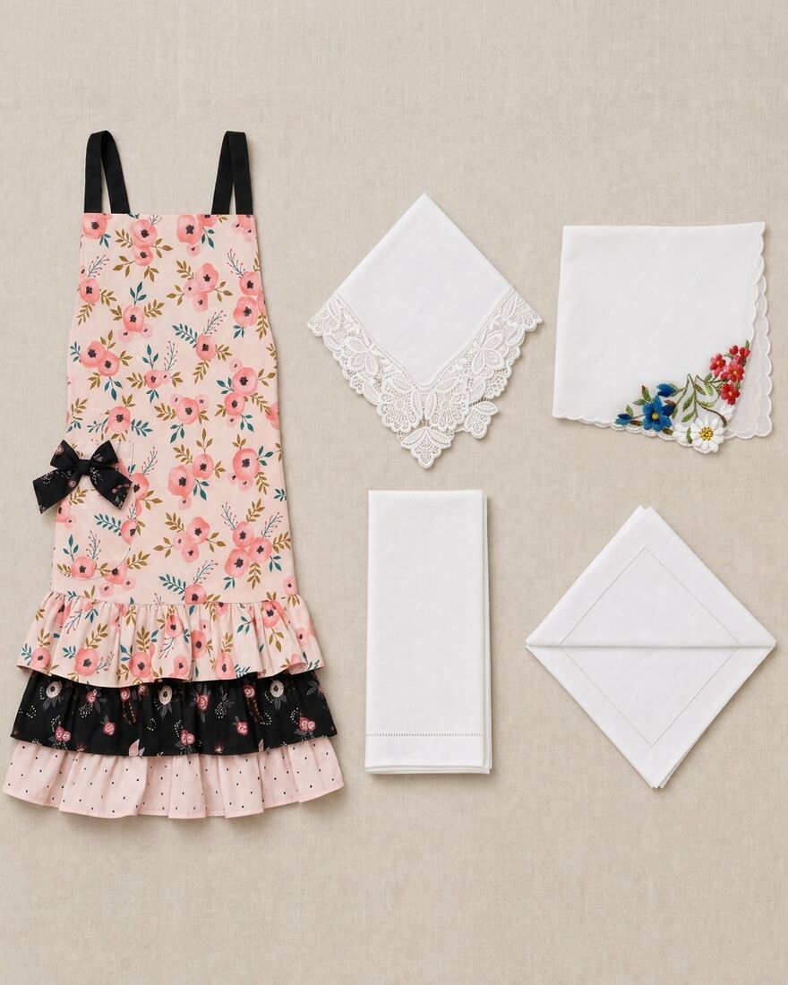 A vintage floral apron, a lace-edged handkerchief, a printed forget-me-not handkerchief, a hemstitched hand towel and a linen napkin laid out on natural linen